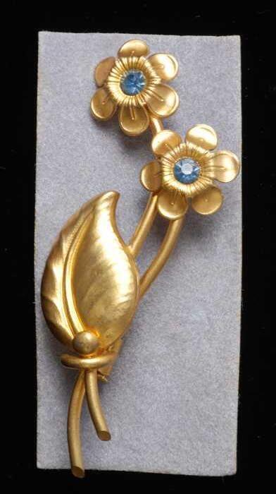 Broche, takje met blad en twee bloemen van goudkleurig metaal met lichtblauwe kunstdiamantjes, op…