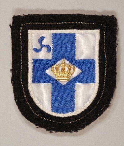 Stoffen badge, op zwart vilten achtergrond blauw kruis op wit fond met kroon en in rechter…