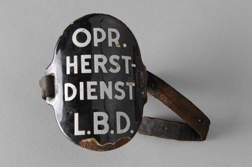 Ovaal zwart geëmailleerd armschildje met "OPR. HERST-DIENST L.B.D.", met (gebroken) leren riempjes…