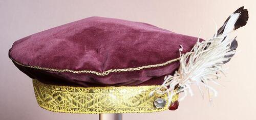 Auberginekleurige fluwelen ronde baret, zwarte pietenpet, met goudlamee band en garnering van twee…