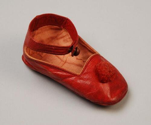 Één rood leren babyschoentje met wollen pompoentje op de voorvoet en enkelbandje met knoopsluiting