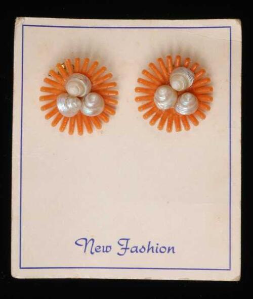 Paar oorbellen, clips met oranje plastic straalvormige bloem met een hart van schelpjes, op karton,…