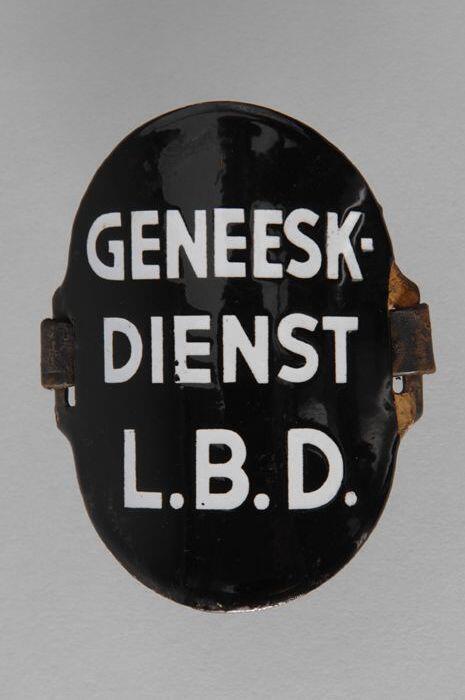 Ovaal zwart geëmailleerd armschildje met "GENEESK. DIENST L.B.D.", met afgescheurde leren riempjes…