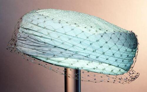 Pillbox, cremekleurige stof met langs rand garnering van pastelkleurige crêpe en beige voile met…