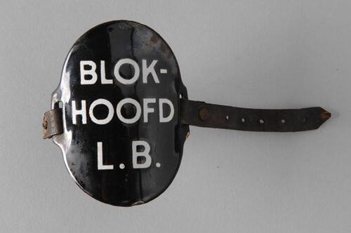 Ovaal zwart geëmailleerd armschildje met "BLOK-HOOFD L.B.", met (afgescheurde) leren riempjes aan…
