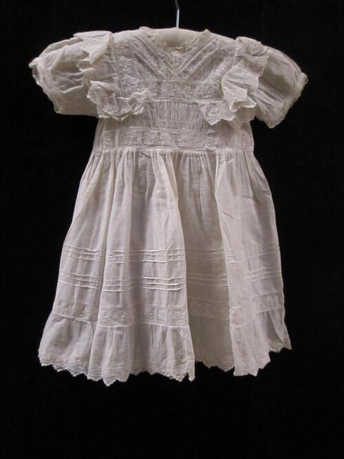 Jurk (kind) van witte broderie, kantrandjes langs hals, dubbele geschulpte volants broderie op de…