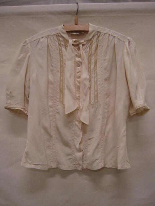 Blouse van crèmekleurige zijde met tussenzetsels van geborduurde tule en een striklint rond de hals