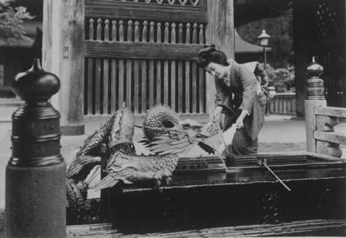 Japanse vrouw haalt met een beker aan een stokje water uit een met een draak opgesierde waterbak in…
