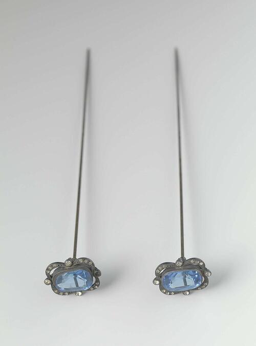Hoedenspeld van staal met een langwerpig ornament van facet geslepen blauw glas gevat in een stalen…