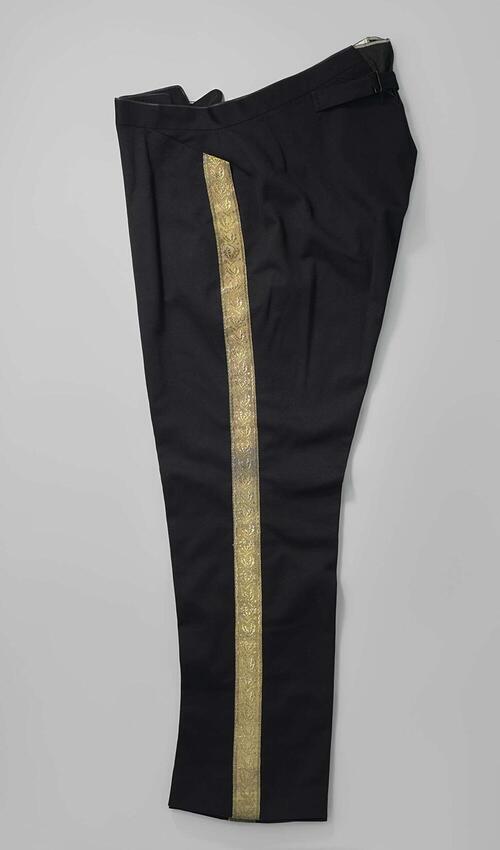 Broek van zwart laken met lange pijpen en opgenaaide tailleband, vanachter met losse banden met een…